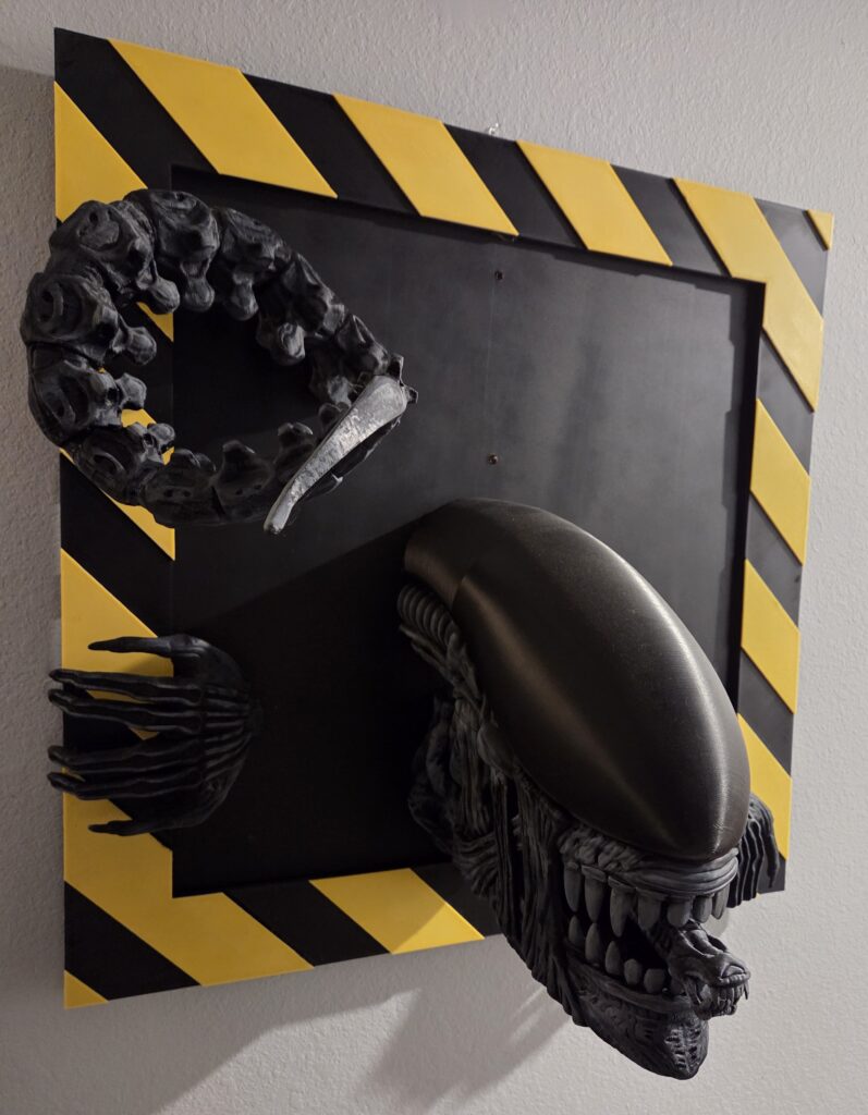 Black Xenomorph alien emerging from a black-and-yellow hazard-striped 3D wall display